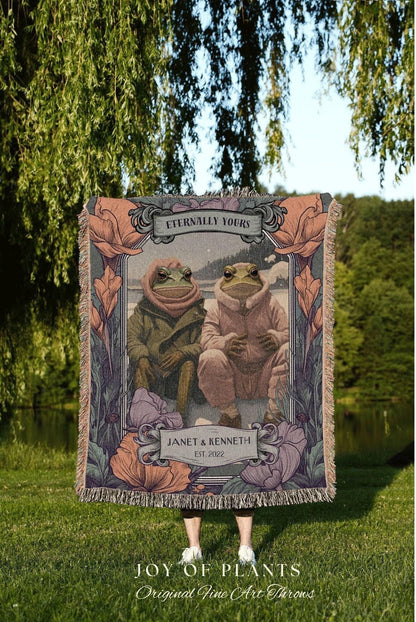 May include: A woven throw blanket featuring a framed illustration of two frogs in hooded outfits, with the text "Eternally Yours" at the top. The blanket has a floral border and the names "Janet & Kenneth" with the year "EST. 2022".