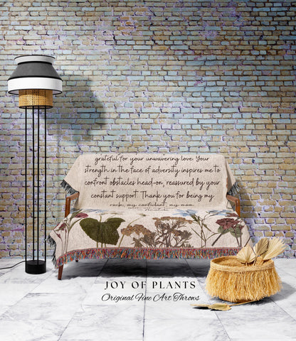 May include: A beige throw blanket with floral designs and the text "grateful for your unwavering love..." is draped over a wooden sofa. A black and white floor lamp stands to the left. A woven basket with decorative fringe and dried plants sits on the floor.