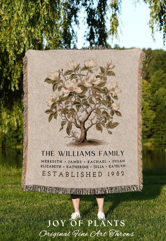 May include: A beige throw blanket featuring a detailed illustration of a magnolia tree with white flowers. The text "THE WILLIAMS FAMILY" and names are printed below the tree, with "ESTABLISHED 1982" beneath. The blanket has fringed edges.
