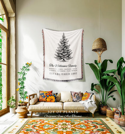 May include: A white throw blanket with a black and white illustration of a Christmas tree, with the text "The Williams Family" and "Established 1982". The blanket is hanging on a wall above a sofa with colorful pillows. The room has a rug with an orange and green pattern.
