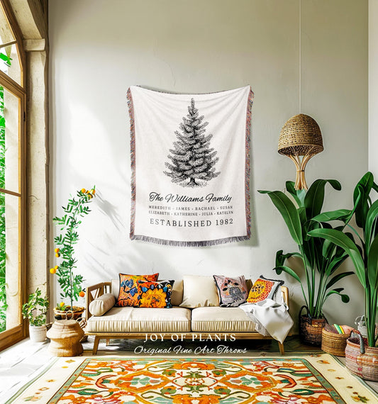 May include: A white throw blanket with a black and white illustration of a Christmas tree, with the text "The Williams Family" and "Established 1982". The blanket is hanging on a wall above a sofa with colorful pillows. The room has a rug with an orange and green pattern.