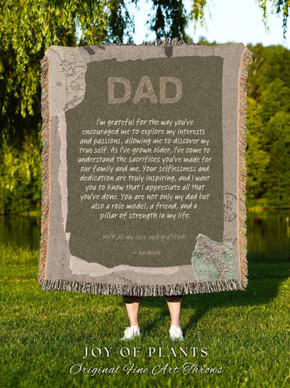 May include: A rectangular throw blanket with the word "DAD" in large, textured letters. The blanket has a message of gratitude and love, with a map design. The blanket is fringed and is held up by a person standing on green grass.