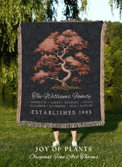 May include: A dark gray throw blanket featuring a detailed illustration of a tree with reddish-brown leaves. The text "The Williams Family" and names are displayed, along with "Established 1982". The blanket has a fringed edge.