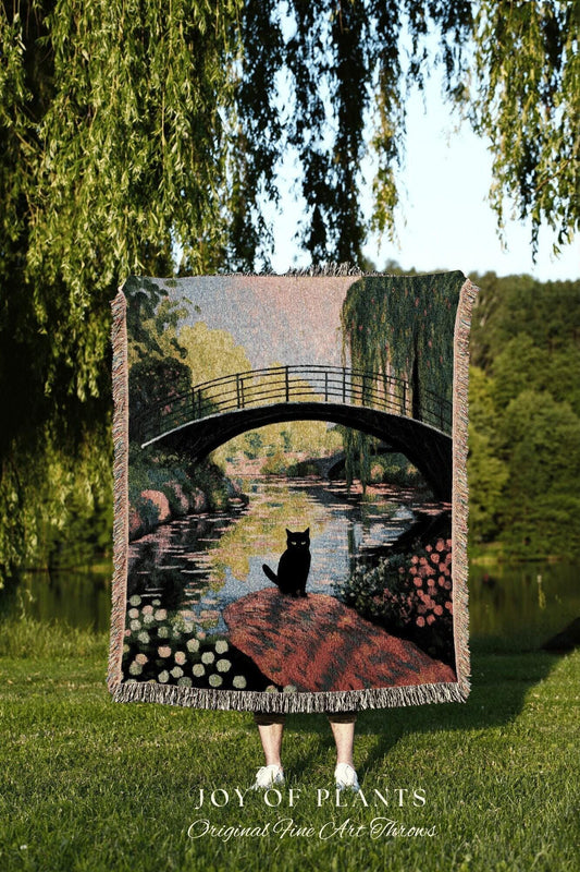 May include: A woven throw blanket featuring a scenic landscape with a black cat. The design depicts a bridge over a river, with trees and flowers. The blanket has a fringed edge and the text "JOY OF PLANTS" and "Original Fine Art Throws".