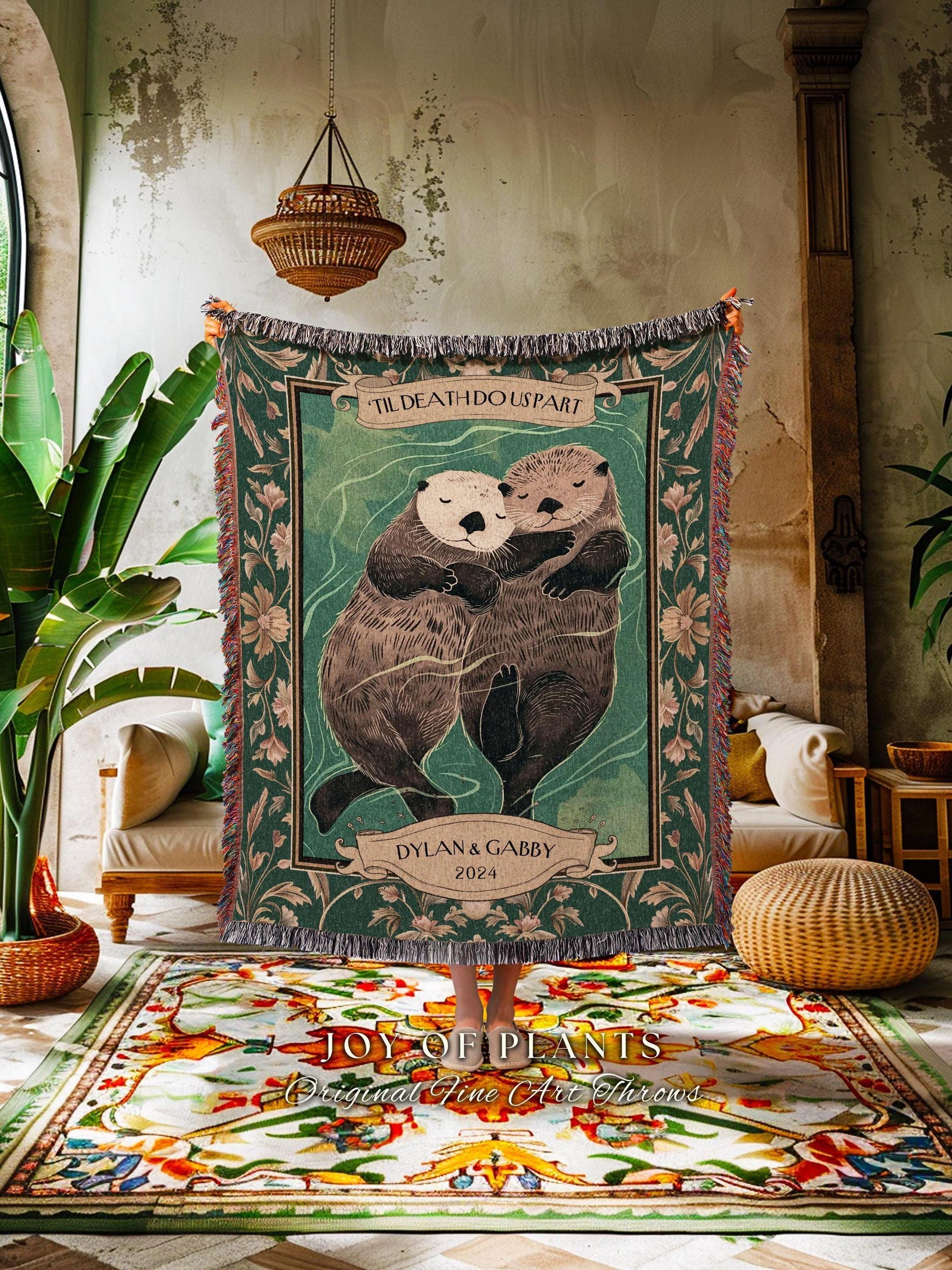 May include: A green woven throw blanket featuring two hugging otters with the text "'TIL DEATH DOUS PART" and "DYLAN & GABBY 2024". The blanket has a fringed edge and floral accents. The blanket is displayed indoors.