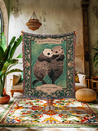 May include: A green woven throw blanket featuring two hugging otters with the text "'TIL DEATH DOUS PART" and "DYLAN & GABBY 2024". The blanket has a fringed edge and floral accents. The blanket is displayed indoors.