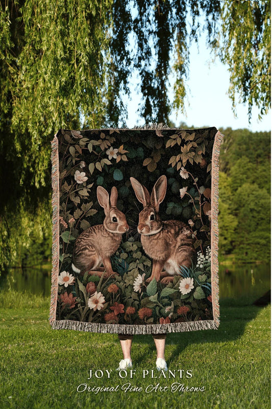 May include: A woven throw blanket featuring a detailed illustration of two brown rabbits amidst a floral and foliage background. The blanket has a dark background with white and red flowers. The text "JOY OF PLANTS Original Fine Art Throws" is visible.