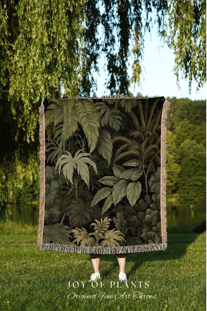 May include: A woven throw blanket featuring a detailed illustration of a lush, green jungle scene. The blanket has a fringed edge and is held up in front of a person. The text "JOY OF PLANTS Original Fine Art Throws" is visible at the bottom.