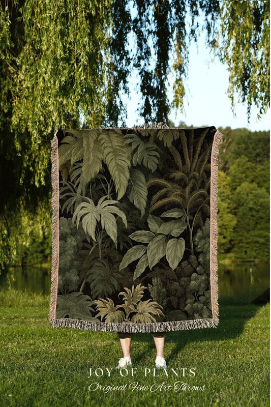May include: A woven throw blanket featuring a detailed illustration of a lush, green jungle scene. The blanket has a fringed edge and is held up in front of a person. The text "JOY OF PLANTS Original Fine Art Throws" is visible at the bottom.