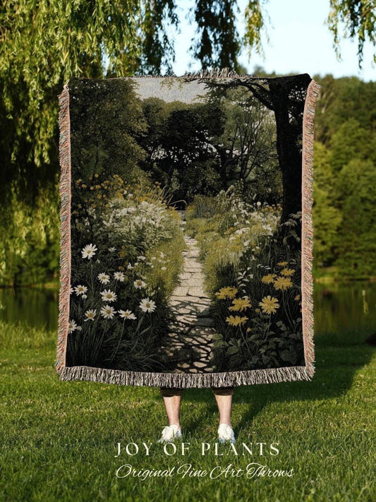 May include: A woven throw blanket featuring a garden path scene with white and yellow flowers. The blanket has a fringed edge and the text "JOY OF PLANTS Original Fine Art Throws" at the bottom. The blanket is held up in front of a person standing on grass.