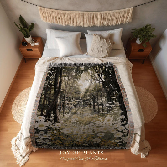May include: A woven throw blanket featuring a forest scene with white flowers and tall trees. The blanket has a cream-colored fringe and is displayed on a bed with white bedding. The text "JOY OF PLANTS Original Fine Art Throws" is visible.
