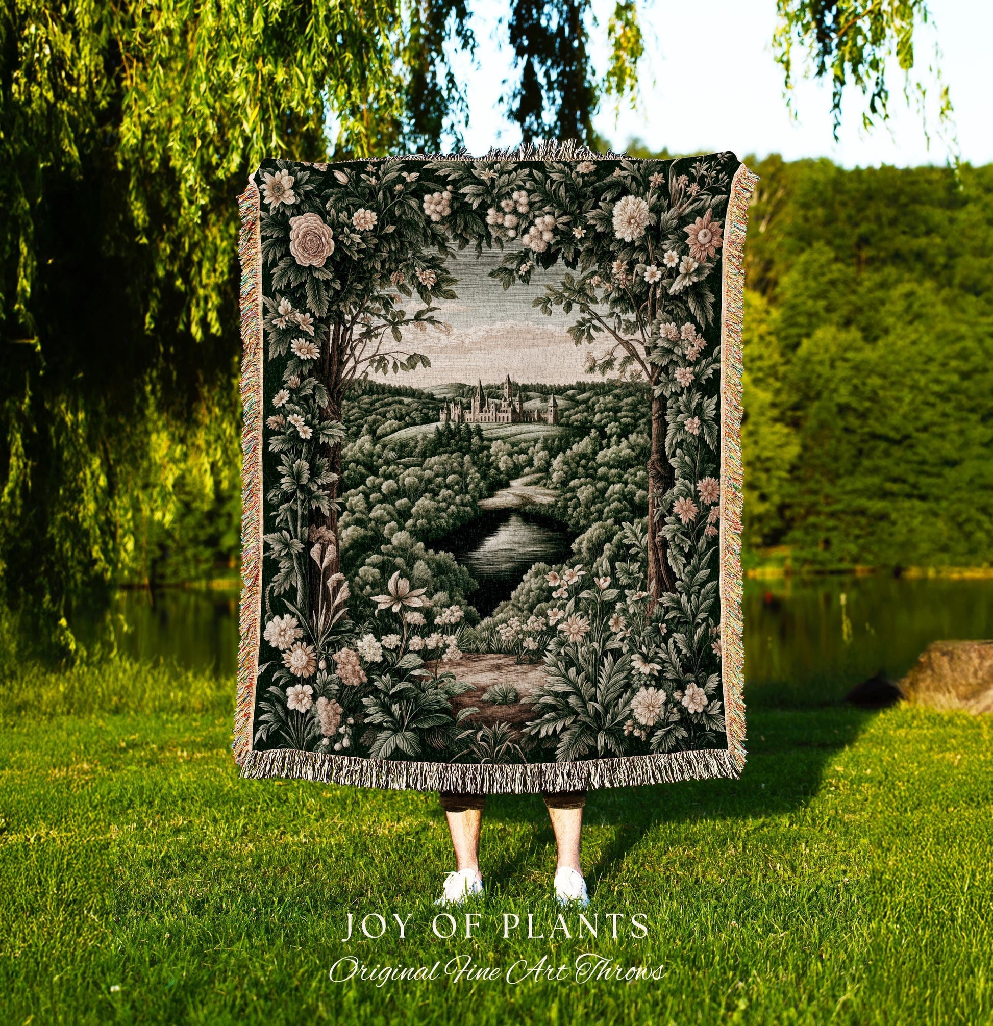 May include: A tapestry throw blanket featuring a landscape scene with a river, trees, and a castle. The blanket has a dark green background with floral and foliage details. The text "JOY OF PLANTS Original Fine Art Throws" is at the bottom.