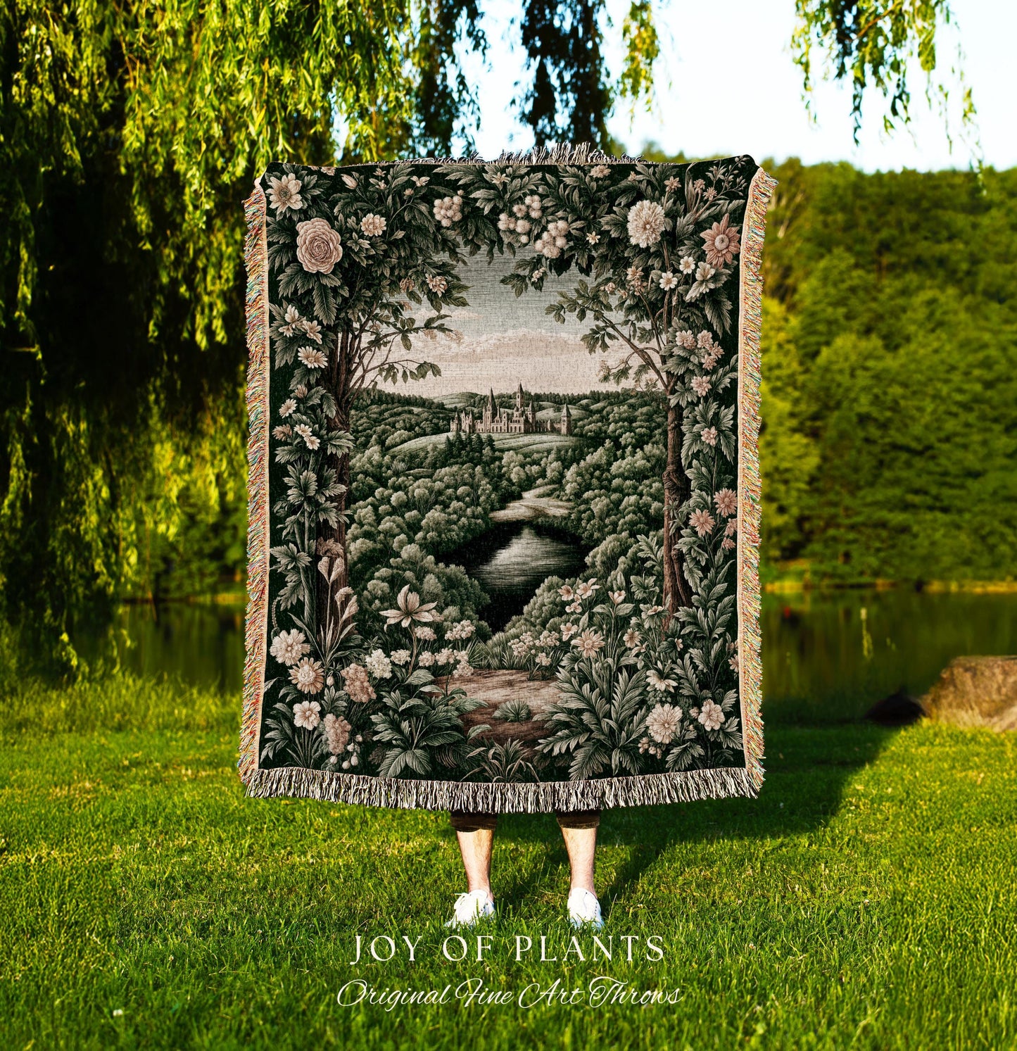 May include: A tapestry throw blanket featuring a landscape scene with a river, trees, and a castle. The blanket has a dark green background with floral and foliage details. The text "JOY OF PLANTS Original Fine Art Throws" is at the bottom.