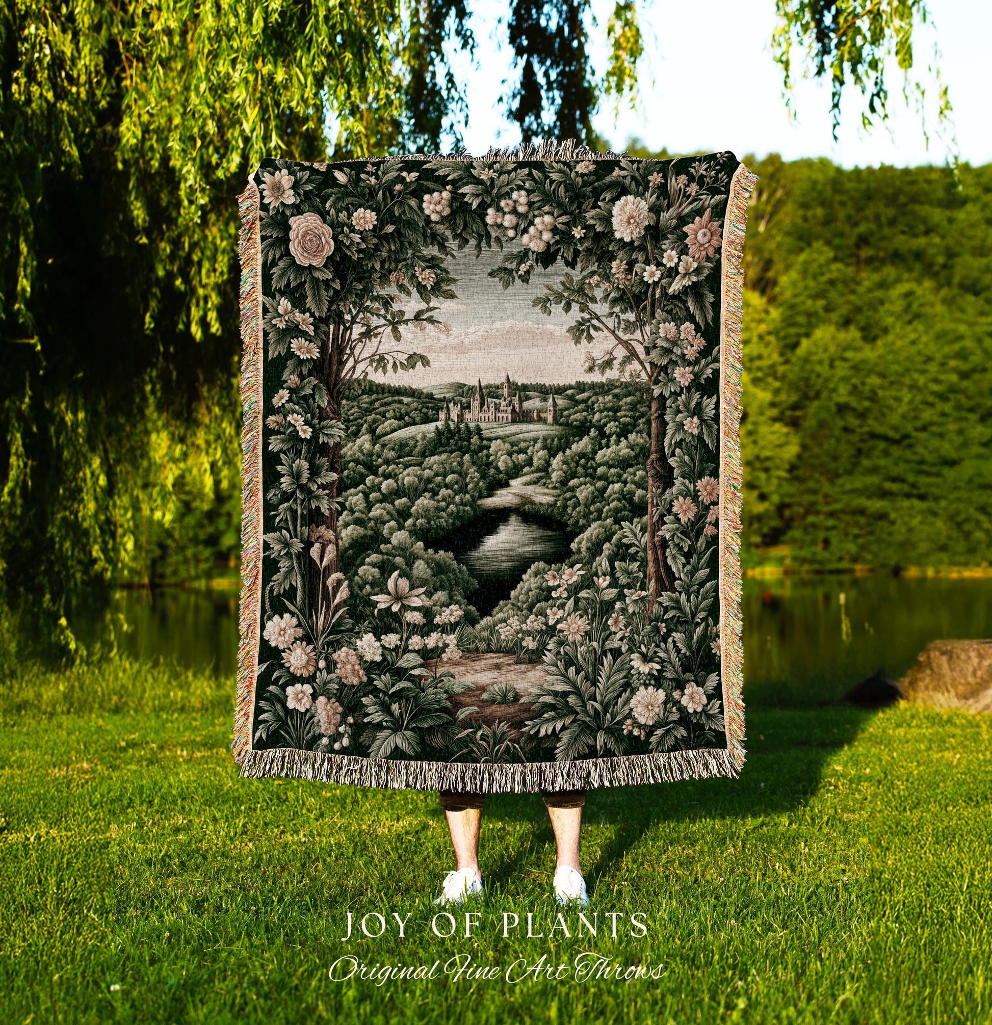 May include: A tapestry throw blanket featuring a landscape scene with a river, trees, and a castle. The blanket has a dark green background with floral and foliage details. The text "JOY OF PLANTS Original Fine Art Throws" is at the bottom.