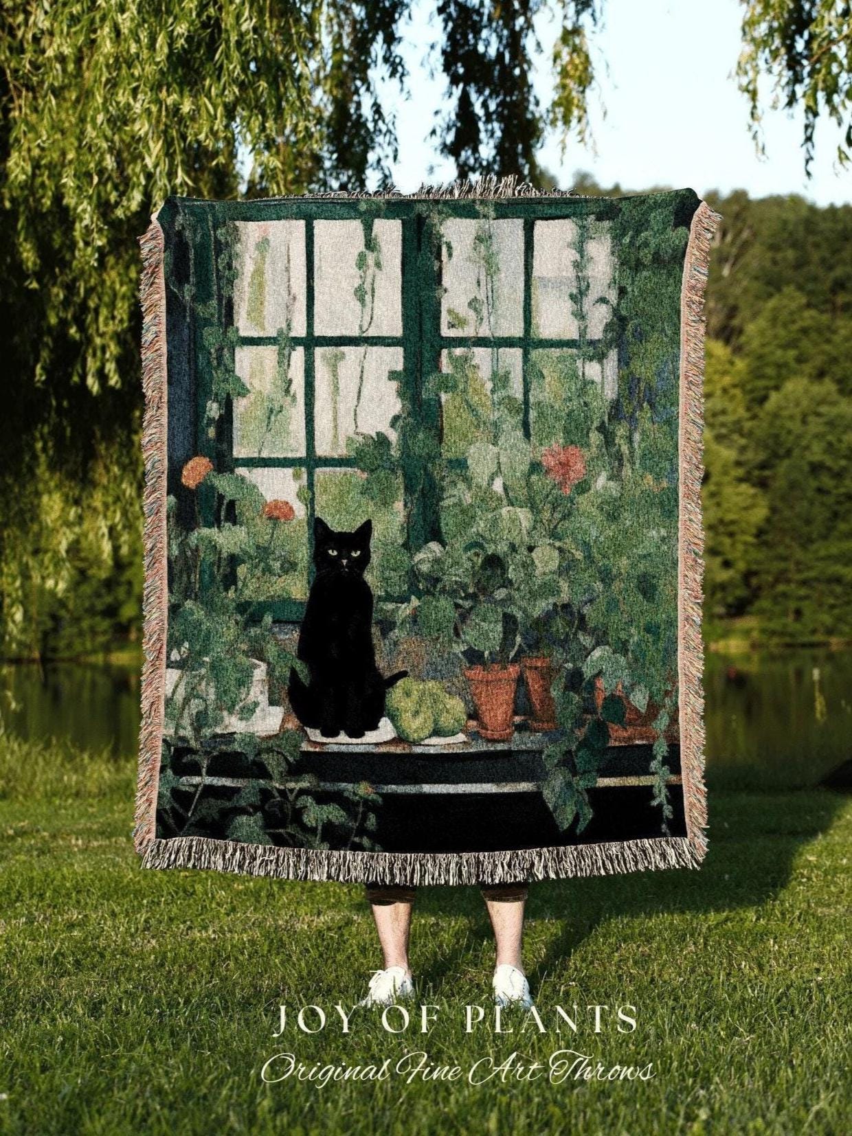 May include: A woven throw blanket featuring a black cat sitting on a windowsill with potted plants. The design includes a green window frame, vines, and the text "JOY OF PLANTS Original Fine Art Throws". The blanket has a fringed edge.
