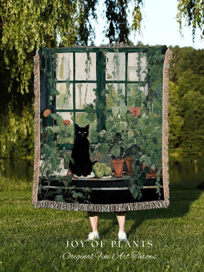 May include: A woven throw blanket featuring a black cat sitting on a windowsill with potted plants. The design includes a green window frame, vines, and the text "JOY OF PLANTS Original Fine Art Throws". The blanket has a fringed edge.