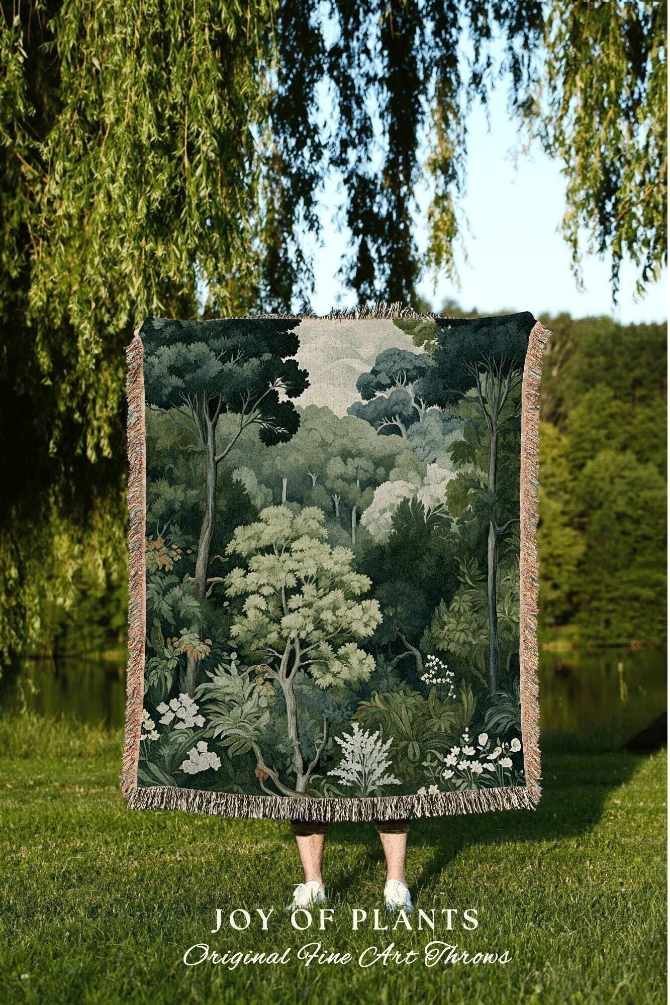 May include: A tapestry throw blanket featuring a detailed forest scene in shades of green, white, and gray. The blanket has a fringed edge and the text "JOY OF PLANTS Original Fine Art Throws" at the bottom. The blanket is held up in front of a person.