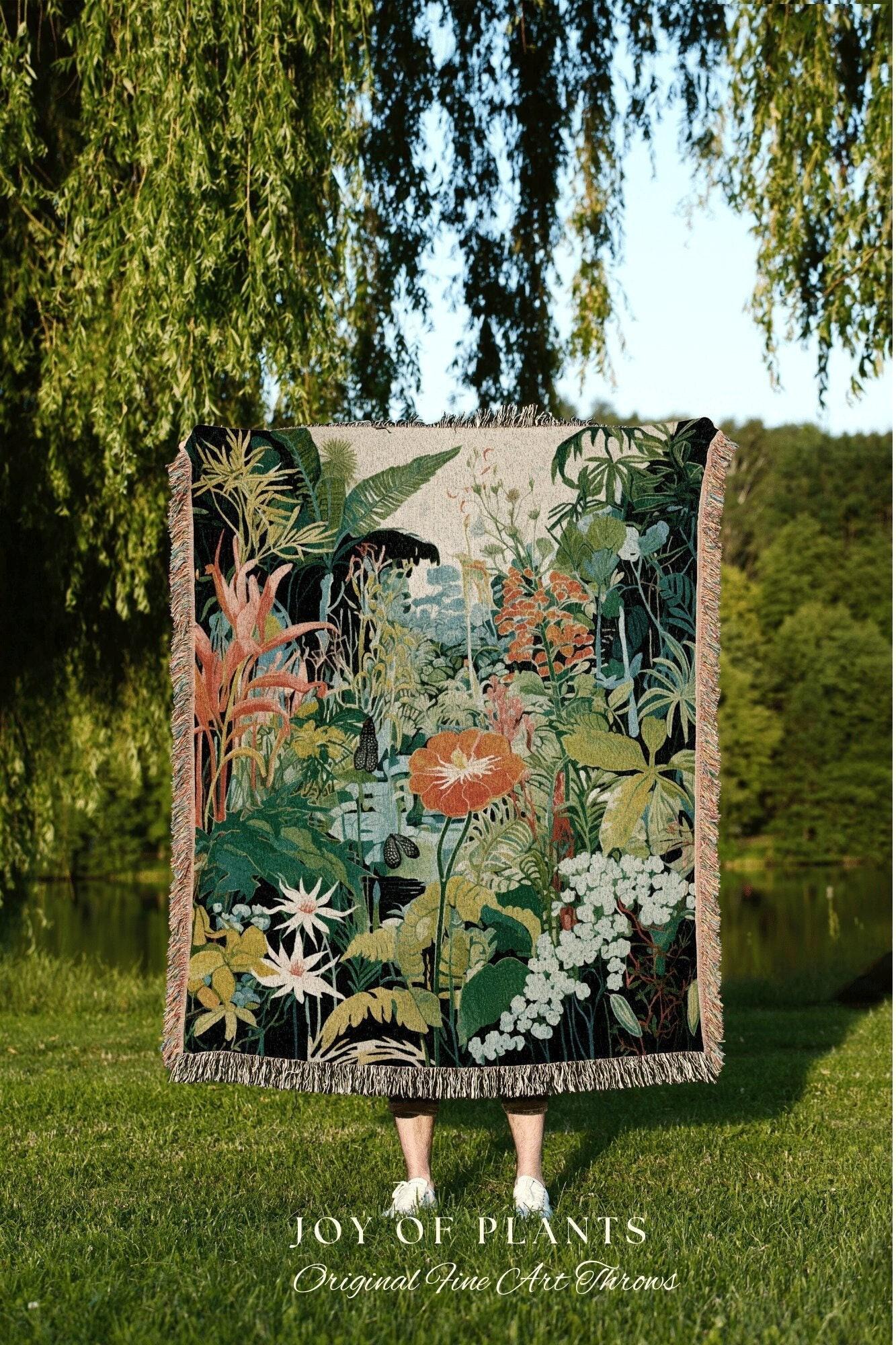 May include: A woven throw blanket featuring a vibrant botanical design with lush greenery, orange flowers, and white blossoms. The blanket has a fringed edge and the text "JOY OF PLANTS Original Fine Art Throws" at the bottom. The blanket is held up in front of a person.