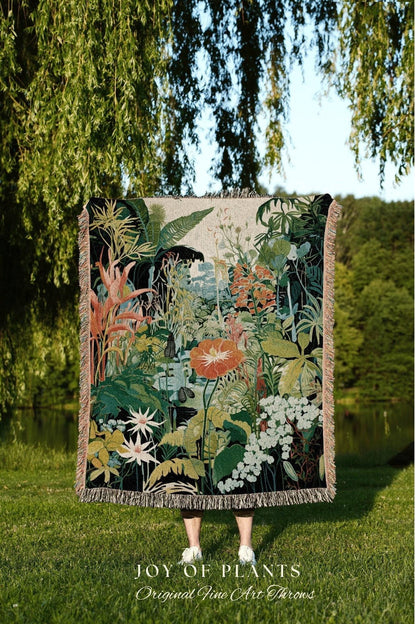 May include: A woven throw blanket featuring a vibrant botanical design with lush greenery, orange flowers, and white blossoms. The blanket has a fringed edge and the text "JOY OF PLANTS Original Fine Art Throws" at the bottom. The blanket is held up in front of a person.