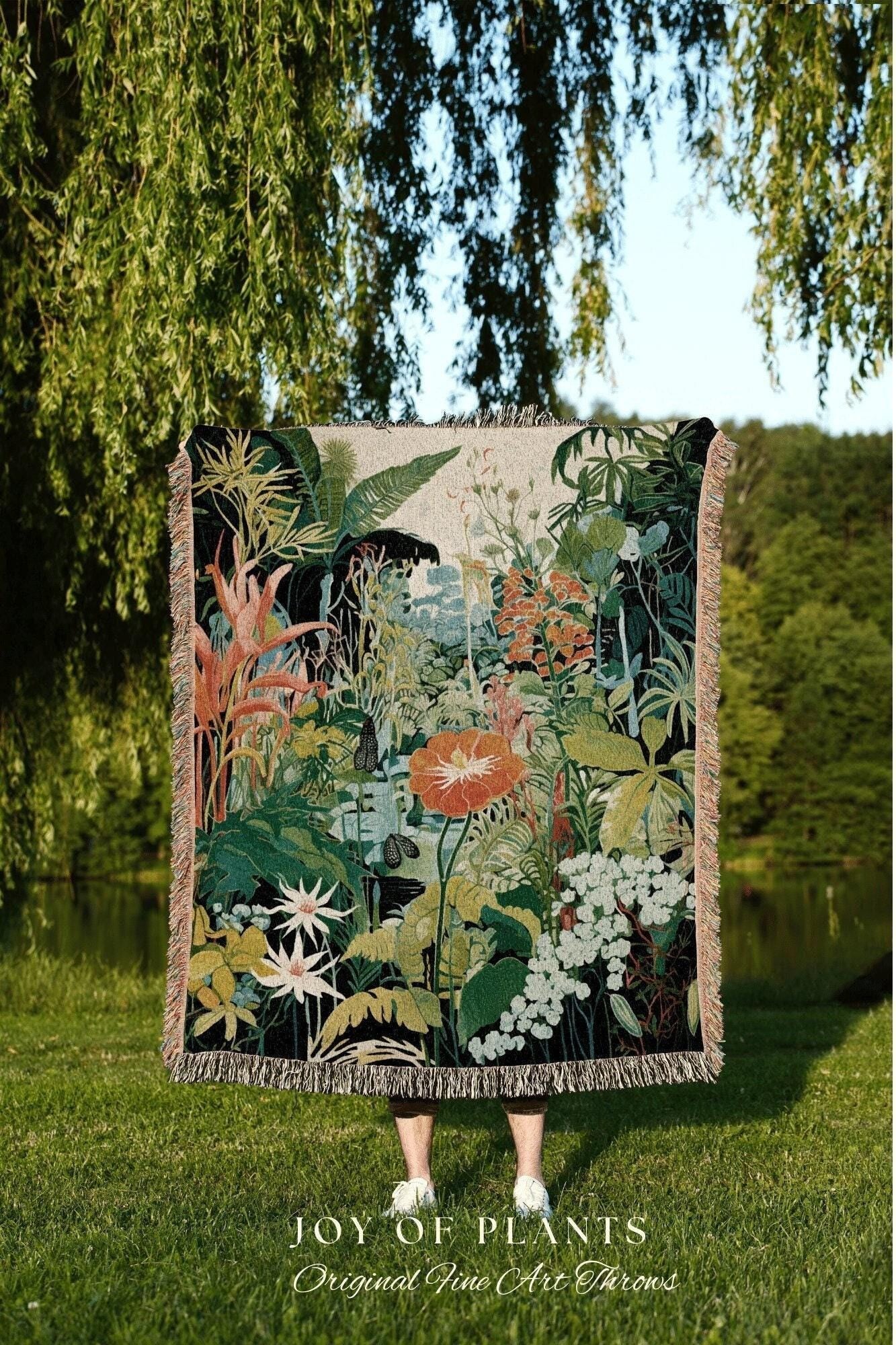 May include: A woven throw blanket featuring a vibrant botanical design with lush greenery, orange flowers, and white blossoms. The blanket has a fringed edge and the text "JOY OF PLANTS Original Fine Art Throws" at the bottom. The blanket is held up in front of a person.
