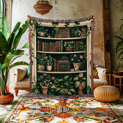 May include: A woven throw blanket featuring a detailed illustration of a bookshelf filled with plants and books. The design includes various potted plants and stacks of books in shades of green, brown, and white. The blanket has a fringed edge.
