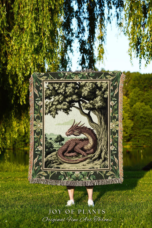 May include: A tapestry throw blanket featuring a dragon resting under a tree. The blanket has a dark green border with floral and leaf patterns. The dragon is brown and red, and the scene is set in a natural landscape. The text "JOY OF PLANTS Original Fine Art Throws" is at the bottom.