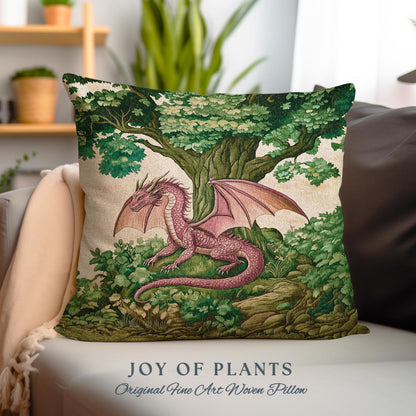 May include: A pink dragon with green wings is nestled in a green forest with a large tree. The dragon is surrounded by green foliage and rocks. The pillow features the text "JOY OF PLANTS Original Fine Art Woven Pillow".