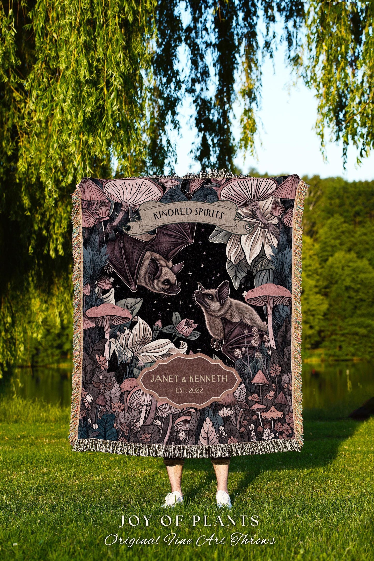 May include: A decorative throw blanket featuring a dark background with bats, mushrooms, and floral elements. The blanket has the text "KINDRED SPIRITS" and "JANET & KENNETH EST 2022". The bottom of the blanket has the text "JOY OF PLANTS Original Fine Art Throws".