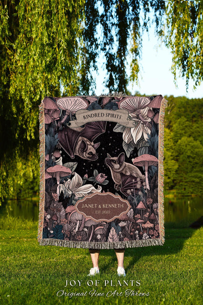 May include: A decorative throw blanket featuring a dark background with bats, mushrooms, and floral elements. The blanket has the text "KINDRED SPIRITS" and "JANET & KENNETH EST 2022". The bottom of the blanket has the text "JOY OF PLANTS Original Fine Art Throws".