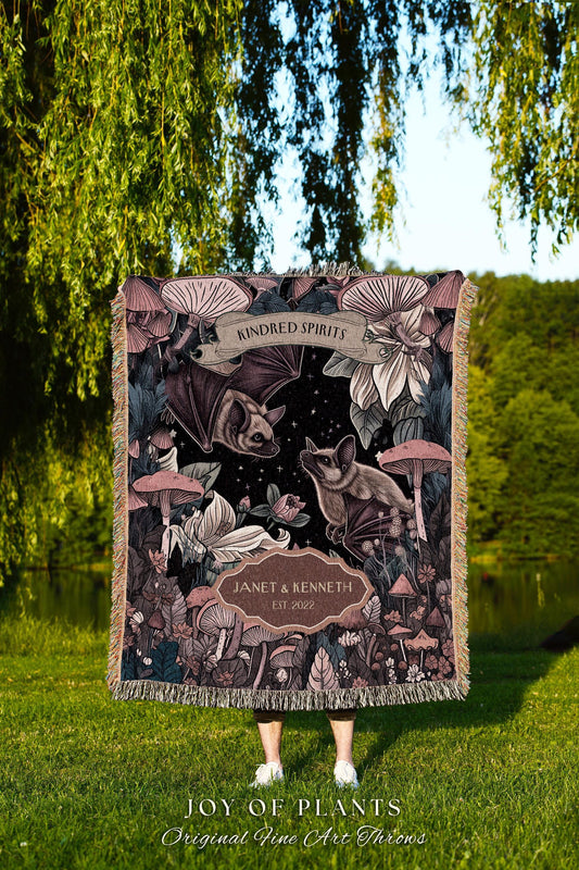 May include: A decorative throw blanket featuring a dark background with bats, mushrooms, and floral elements. The blanket has the text "KINDRED SPIRITS" and "JANET & KENNETH EST 2022". The bottom of the blanket has the text "JOY OF PLANTS Original Fine Art Throws".