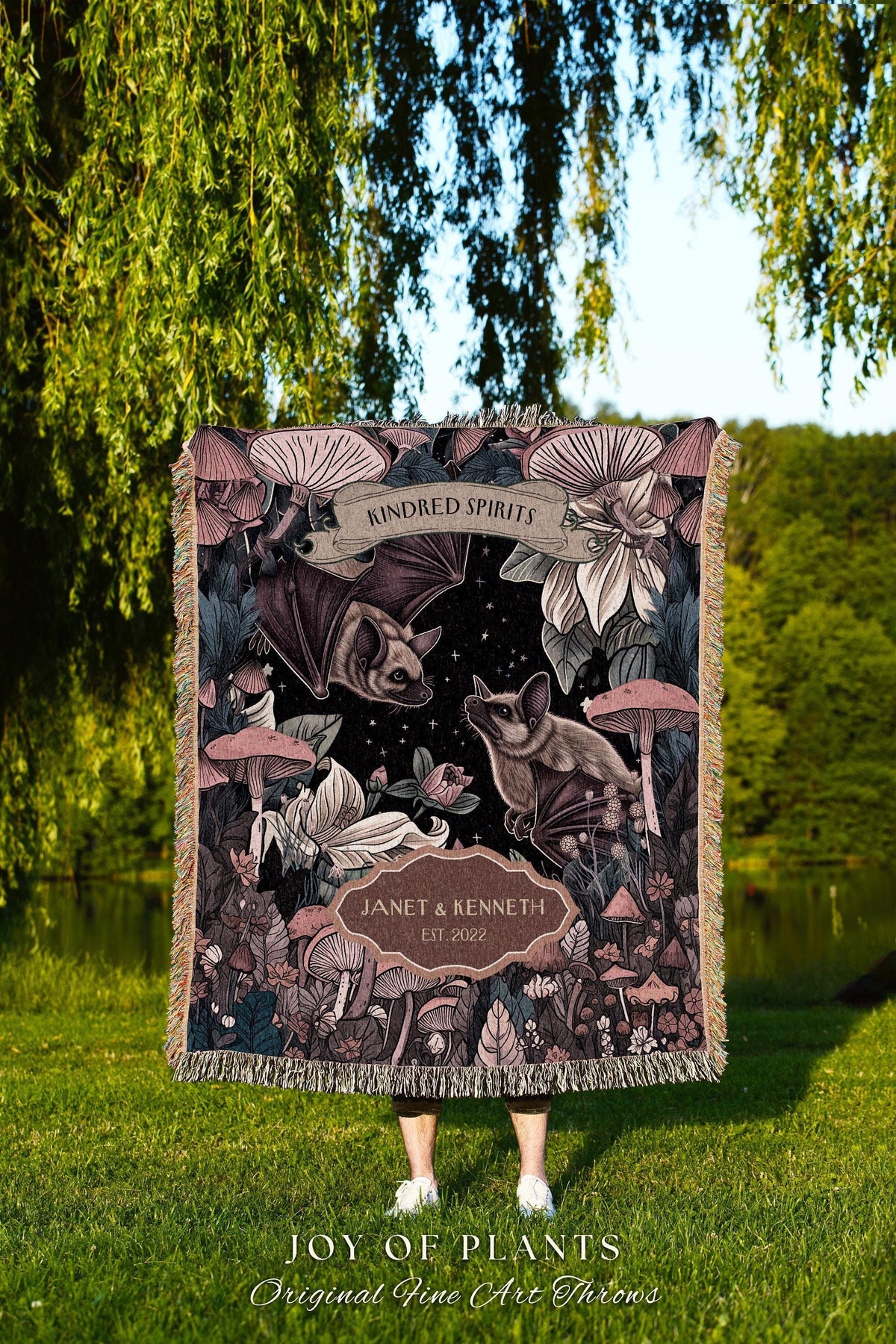 May include: A decorative throw blanket featuring a dark background with bats, mushrooms, and floral elements. The blanket has the text "KINDRED SPIRITS" and "JANET & KENNETH EST 2022". The bottom of the blanket has the text "JOY OF PLANTS Original Fine Art Throws".