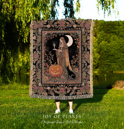 May include: A decorative tapestry throw blanket featuring a mystical scene with a woman, a jack-o'-lantern, and a black cat under a crescent moon. The blanket has a black background with floral and botanical patterns and fringe.