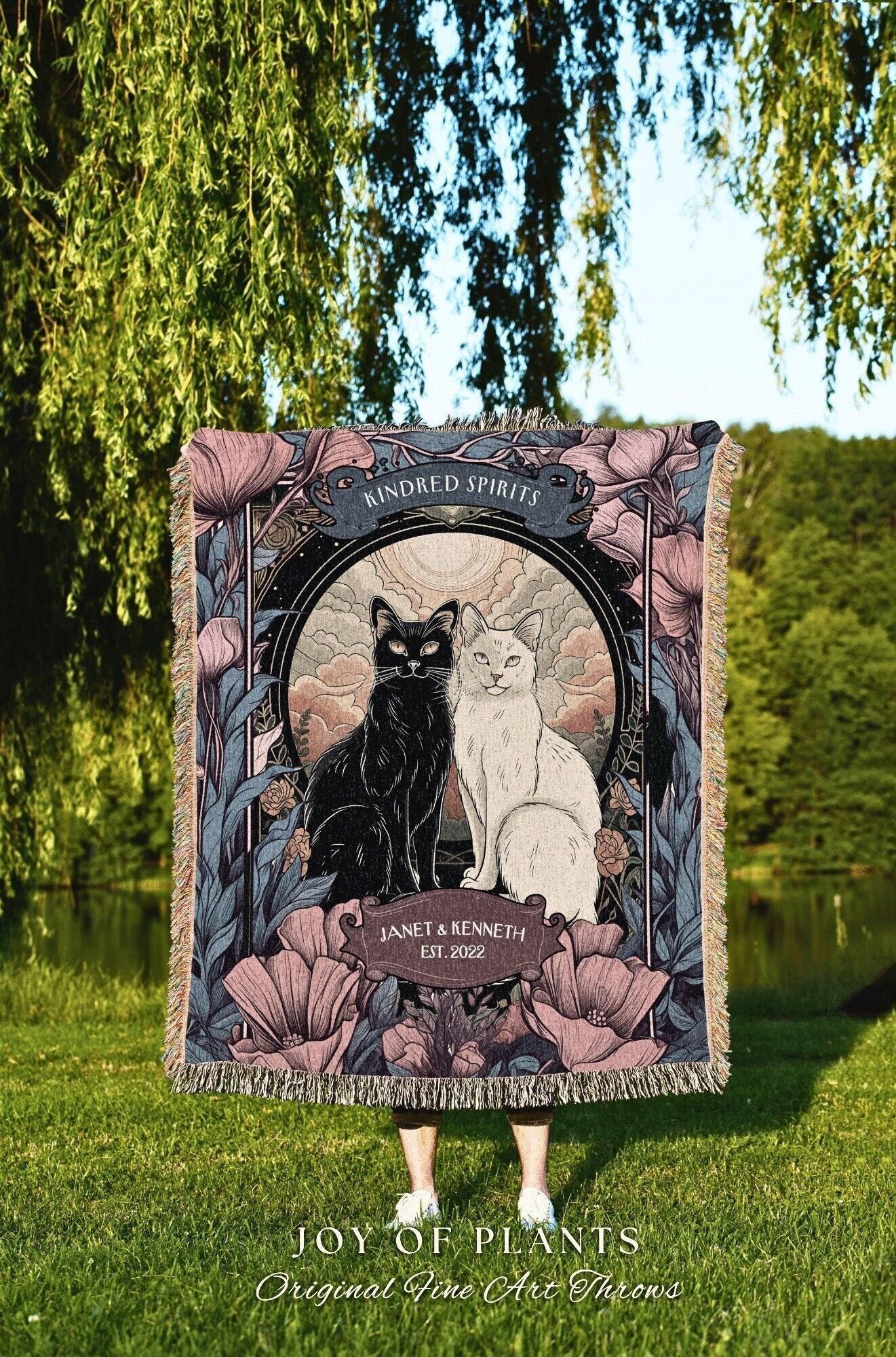 May include: A woven throw blanket featuring a black and white cat illustration within a circular frame, surrounded by floral elements. The text "Kindred Spirits" and "Janet & Kenneth EST. 2022" are displayed. The blanket is held outdoors, with the text "JOY OF PLANTS Original Fine Art Throws" at the bottom.
