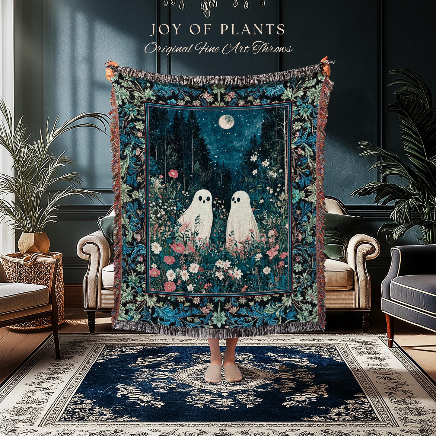 May include: A decorative throw blanket featuring a whimsical forest scene with two white ghost figures, a full moon, and floral accents. The blanket has a dark blue background with a floral border and fringe. Text on the top reads "JOY of PLANTS Original Fine Art Throws."