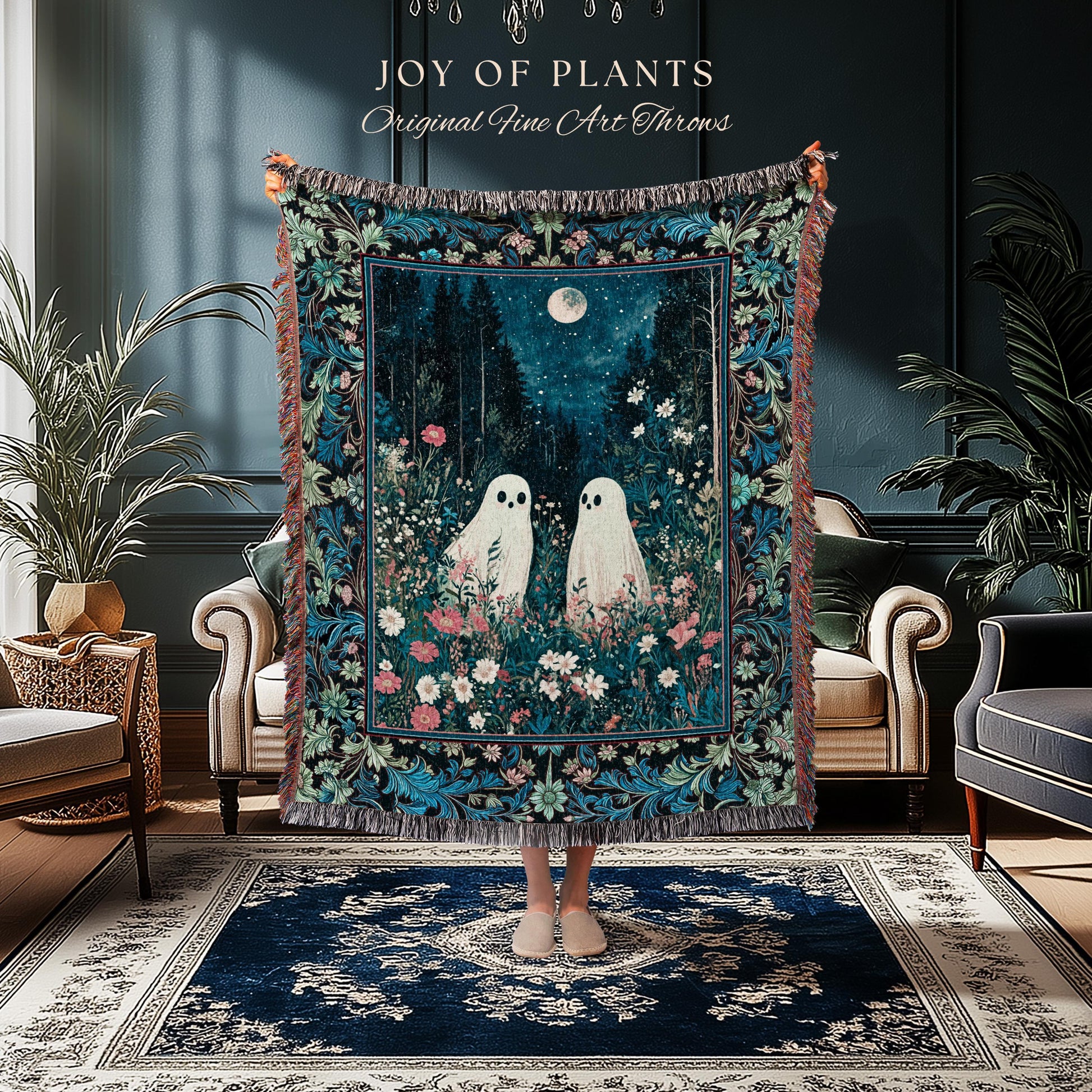May include: A decorative throw blanket featuring a whimsical forest scene with two white ghost figures, a full moon, and floral accents. The blanket has a dark blue background with a floral border and fringe. Text on the top reads "JOY of PLANTS Original Fine Art Throws."