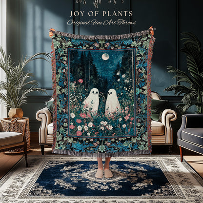 May include: A decorative throw blanket featuring a whimsical forest scene with two white ghost figures, a full moon, and floral accents. The blanket has a dark blue background with a floral border and fringe. Text on the top reads "JOY of PLANTS Original Fine Art Throws."