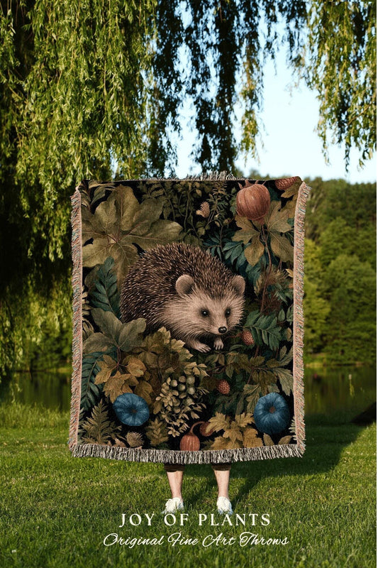 May include: A tapestry throw blanket featuring a detailed illustration of a hedgehog surrounded by foliage, including leaves, berries, and pumpkins. The blanket has a fringed edge and the text "JOY OF PLANTS Original Fine Art Throws".