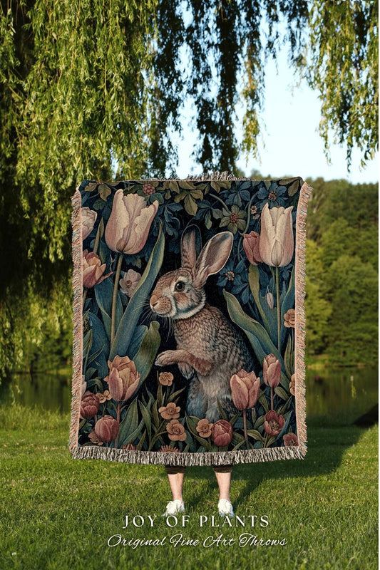 May include: A decorative throw blanket featuring a rabbit amidst tulips and foliage in shades of pink, green, and brown. The blanket has a fringed edge and the text "JOY OF PLANTS Original Fine Art Throws".