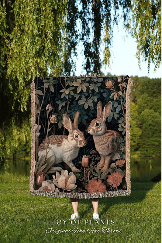 May include: A decorative throw blanket featuring two rabbits in a garden setting. The artwork has a dark background with floral and foliage elements. The blanket has a fringed edge and the text "JOY OF PLANTS Original Fine Art Throws".