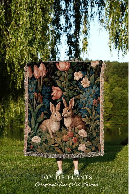 May include: A tapestry throw blanket featuring a floral and fauna design with two rabbits. The throw has a dark background with pink and white flowers, and green foliage. The text "JOY OF PLANTS Original Fine Art Throws" is at the bottom.
