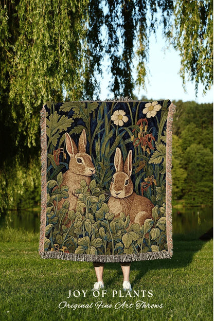 May include: A tapestry throw blanket featuring two brown rabbits amidst lush green foliage and white flowers against a dark blue background. The blanket has a fringed edge and the text "JOY OF PLANTS Original Fine Art Throws".