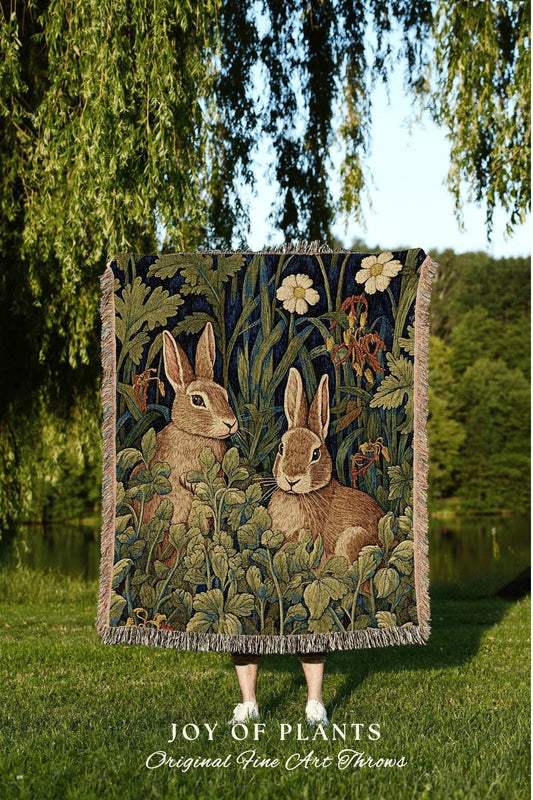 May include: A tapestry throw blanket featuring two brown rabbits amidst lush green foliage and white flowers against a dark blue background. The blanket has a fringed edge and the text "JOY OF PLANTS Original Fine Art Throws".