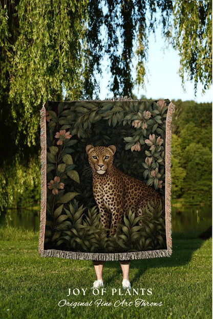 May include: A woven throw blanket featuring a detailed illustration of a leopard in a lush, green jungle setting with pink flowers. The blanket has a fringed edge and the text "JOY OF PLANTS Original Fine Art Throws".