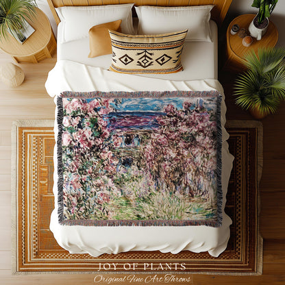May include: A woven throw blanket with a floral painting design in shades of pink, green, and blue. The blanket has a fringed edge and is displayed on a white bed. The text "JOY OF PLANTS Original Fine Art Throws" is at the bottom.