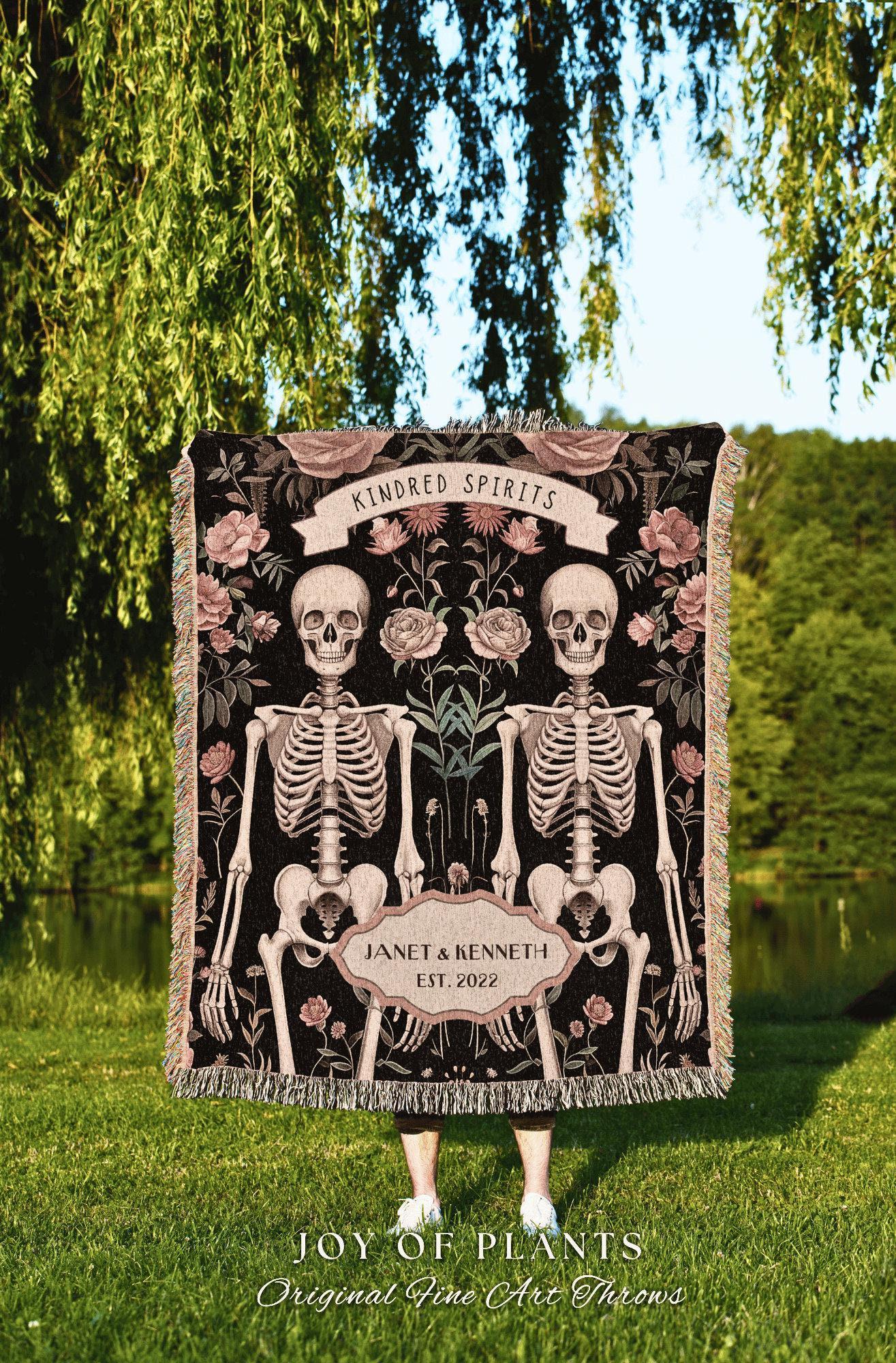 May include: A black throw blanket featuring two skeletons, floral accents, and the text "Kindred Spirits." The blanket includes the names "Janet & Kenneth" and the year "2022." The bottom of the blanket reads "JOY OF PLANTS" and "Original Fine Art Throws."