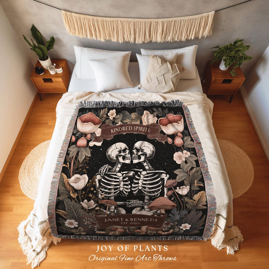 May include: A black throw blanket featuring a skeleton couple embracing, surrounded by floral and mushroom illustrations. The blanket has a fringed edge and the text "KINDRED SPIRITS" and "JANET & KENNETH EST. 2022". The blanket is on a bed with white pillows.