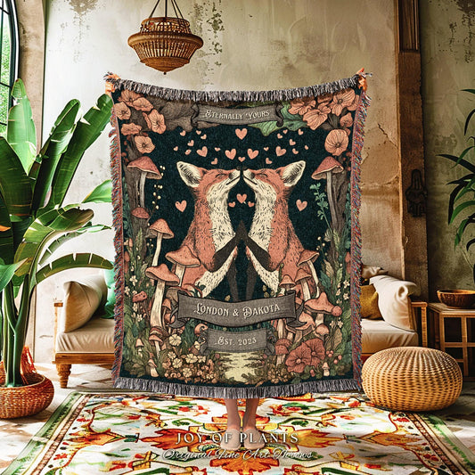 May include: A woven throw blanket featuring a forest scene with two kissing foxes, mushrooms, and floral elements. The blanket has a dark background with the text "Eternally Yours", "London & Dakota", and "Est. 2023". The blanket has a fringed edge.