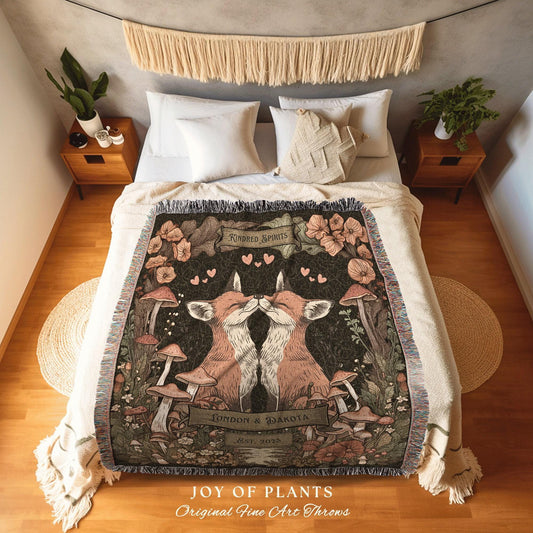 May include: A woven throw blanket featuring a design of two kissing foxes surrounded by mushrooms and flowers. The blanket has a dark background with the words "Kindred Spirits" and "London & Dakota Est. 2023". The bottom of the blanket reads "JOY OF PLANTS Original Fine Art Throws".