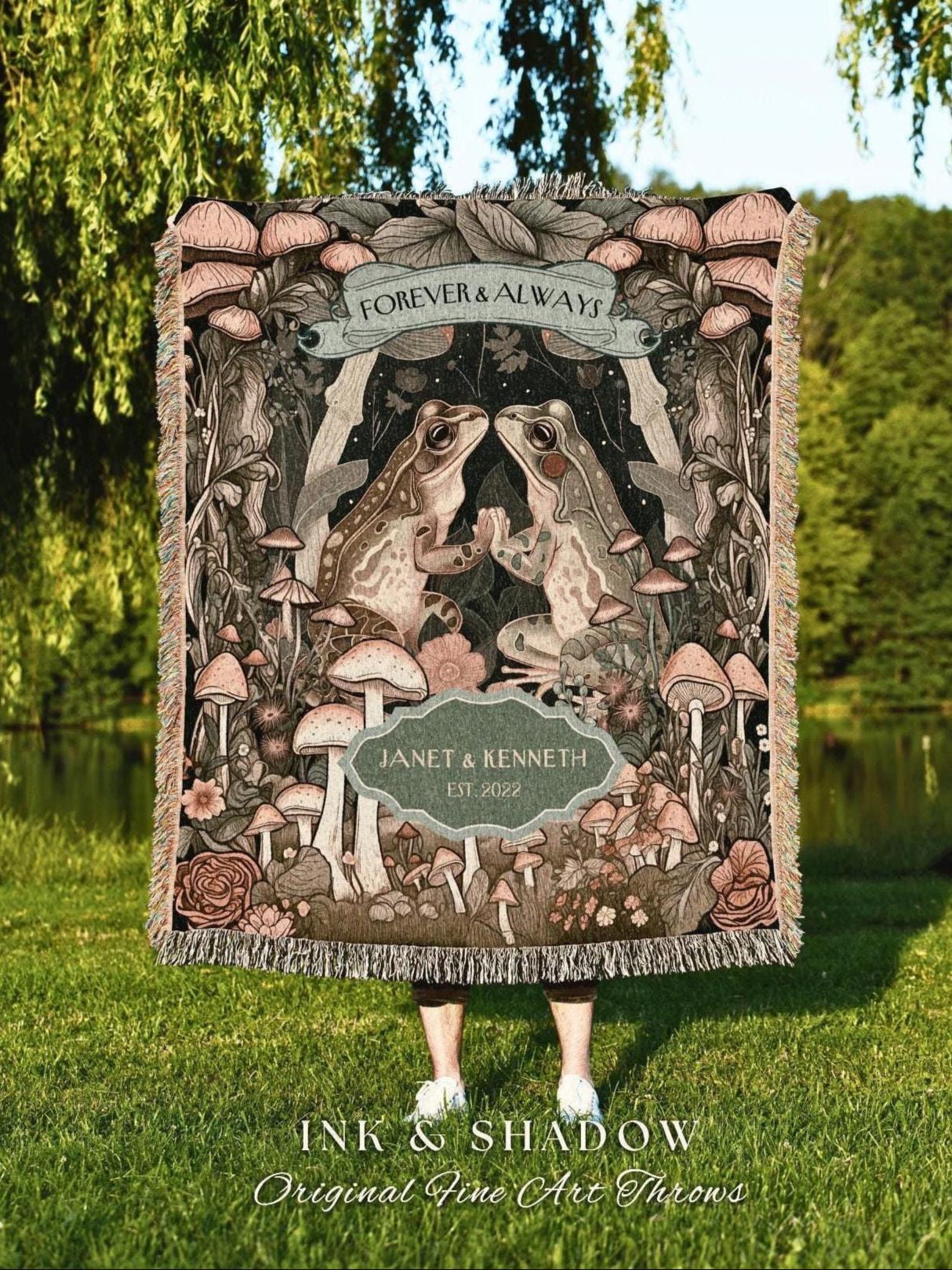 May include: A woven throw blanket featuring a whimsical forest scene with two frogs, mushrooms, and floral elements. The blanket has the text "FOREVER & ALWAYS" and "JANET & KENNETH EST. 2022". The bottom of the blanket reads "INK & SHADOW Original Fine Art Throws".