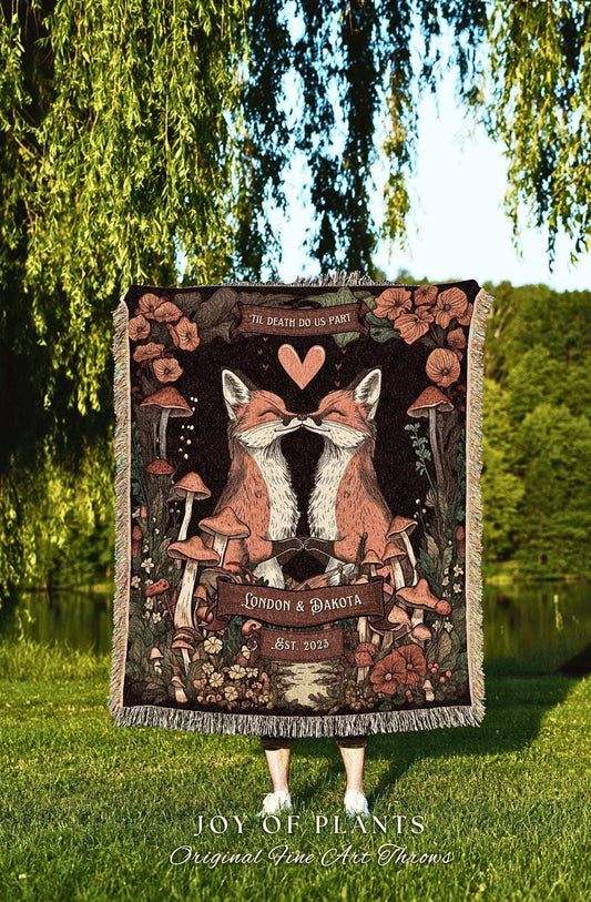 May include: A decorative throw blanket featuring two kissing foxes with a heart above them, surrounded by mushrooms and flowers. The blanket has the text "TIL DEATH DO US PART", "LONDON & DAKOTA", and "EST. 2023". The bottom of the blanket reads "JOY OF PLANTS Original Fine Art Throws".